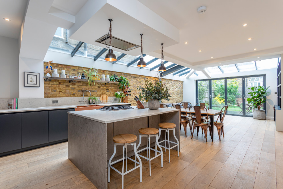 Contemporary galley kitchen in Sussex with a farmhouse sink, flat-panel cabinets, grey cabinets, brown splashback, brick splashback, stainless steel appliances, medium hardwood floors, with island, brown floor and white benchtop.