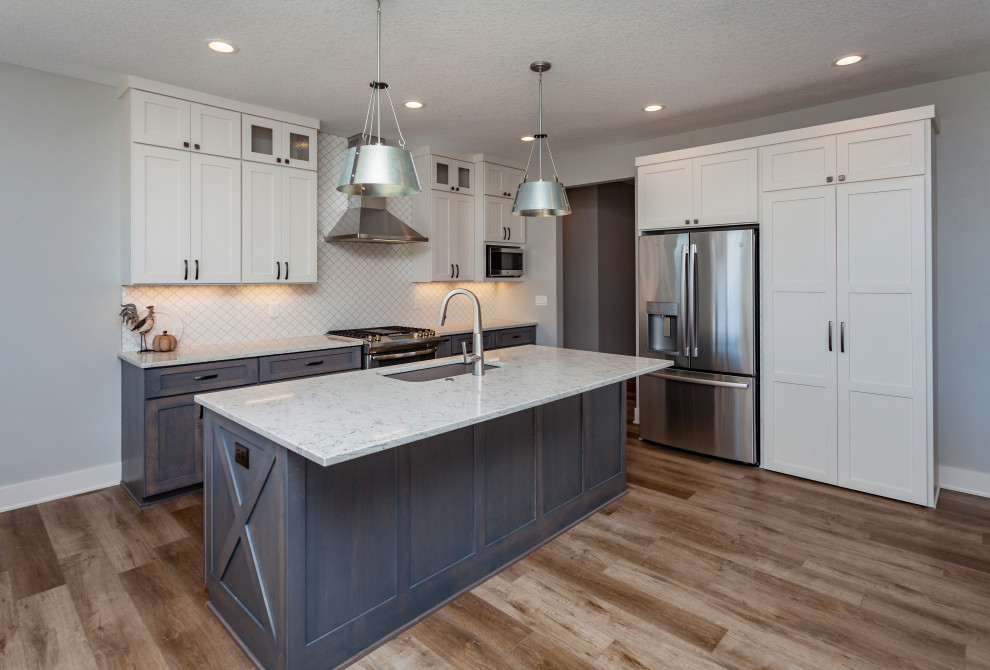 Mid-sized transitional l-shaped light wood floor and brown floor open concept kitchen photo with an undermount sink, shaker cabinets, white cabinets, quartz countertops, white backsplash, ceramic backsplash, stainless steel appliances, an island and white countertops