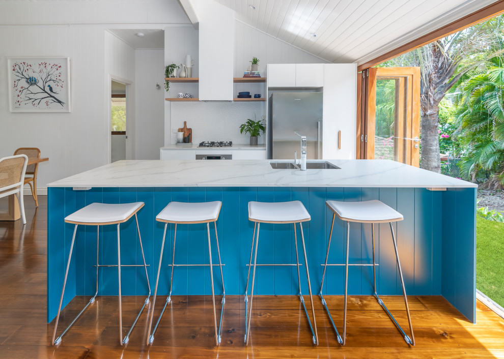 Mid-sized contemporary galley open plan kitchen in Brisbane with dark hardwood floors, a double-bowl sink, blue cabinets, solid surface benchtops, white splashback, mosaic tile splashback, stainless steel appliances, with island and white benchtop.
