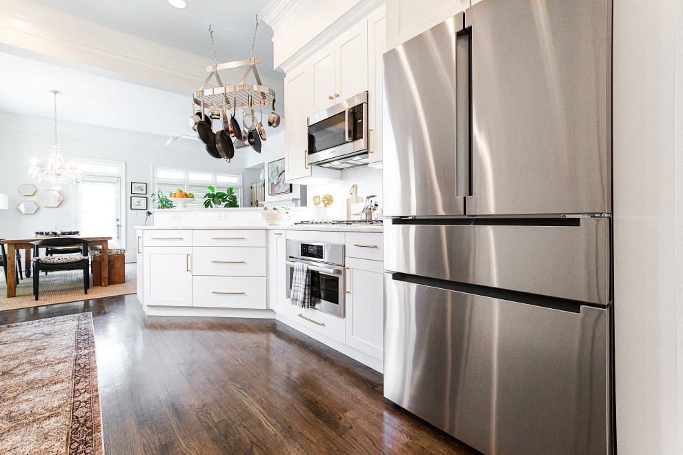 Mid-sized eclectic galley open plan kitchen in Atlanta with an undermount sink, shaker cabinets, white cabinets, quartzite benchtops, white splashback, porcelain splashback, stainless steel appliances, medium hardwood floors, a peninsula, brown floor and white benchtop.