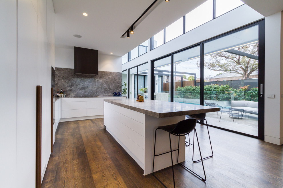 Inspiration for a large modern l-shaped open plan kitchen in Melbourne with an undermount sink, flat-panel cabinets, white cabinets, grey splashback, marble splashback, black appliances, medium hardwood floors, with island, brown floor, grey benchtop and limestone benchtops.