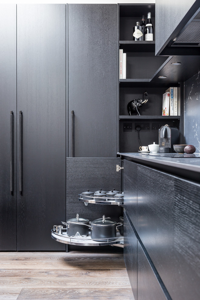 Inspiration for a mid-sized industrial l-shaped open plan kitchen in London with an integrated sink, flat-panel cabinets, black cabinets, solid surface benchtops, black splashback, stone slab splashback, black appliances, medium hardwood floors, with island, beige floor and black benchtop.