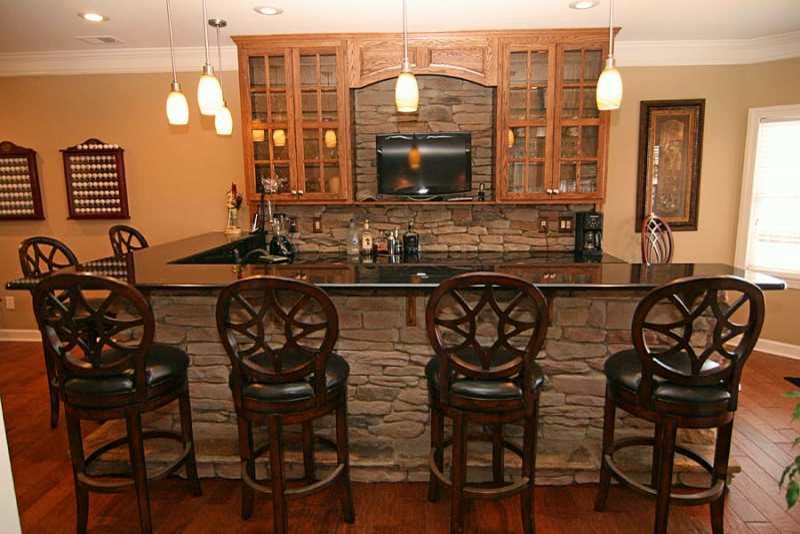 Large transitional l-shaped home bar in Denver with glass-front cabinets, medium wood cabinets, granite benchtops, beige splashback, stone slab splashback and medium hardwood floors.