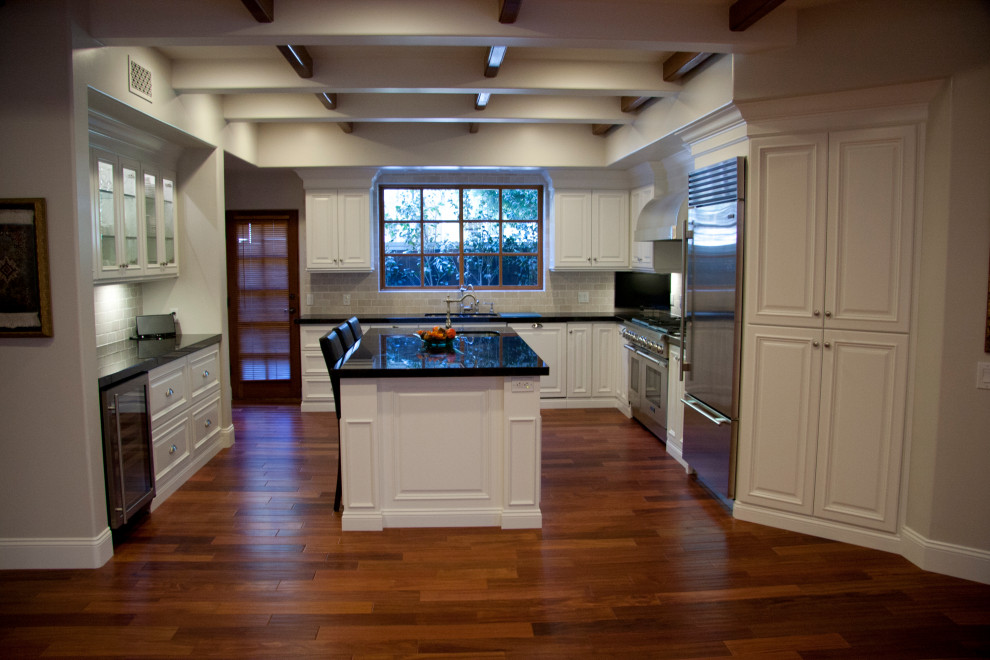 Photo of a large traditional u-shaped eat-in kitchen in Orange County with an undermount sink, raised-panel cabinets, white cabinets, granite benchtops, beige splashback, travertine splashback, stainless steel appliances, medium hardwood floors, with island, red floor and black benchtop.