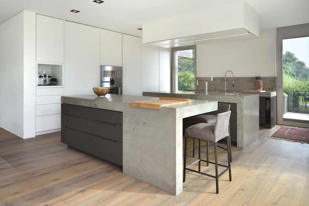 Inspiration for a modern open plan kitchen in Munich with flat-panel cabinets, grey cabinets, concrete benchtops, multiple islands, grey splashback, stainless steel appliances and light hardwood floors.