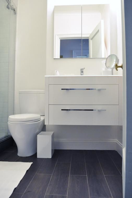 Photo of a small modern 3/4 bathroom in New York with a wall-mount sink, flat-panel cabinets, white cabinets, a corner shower, white tile, subway tile, white walls, a two-piece toilet and vinyl floors.