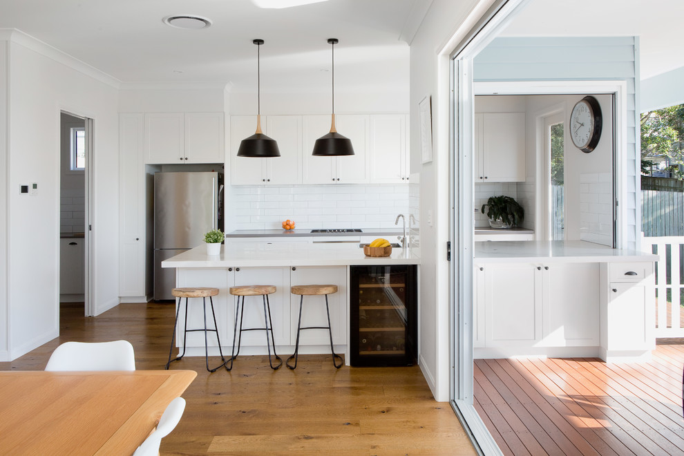 Mid-sized contemporary galley open plan kitchen in Brisbane with white cabinets, cement tile splashback, stainless steel appliances, medium hardwood floors, with island, a drop-in sink, quartz benchtops, white splashback, brown floor and white benchtop.