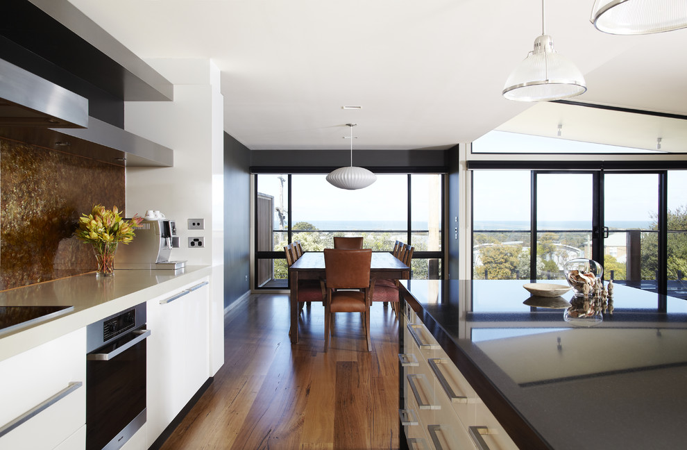 This is an example of a mid-sized beach style single-wall eat-in kitchen in Melbourne with flat-panel cabinets, white cabinets, brown splashback, granite benchtops, stone slab splashback, stainless steel appliances, light hardwood floors and with island.