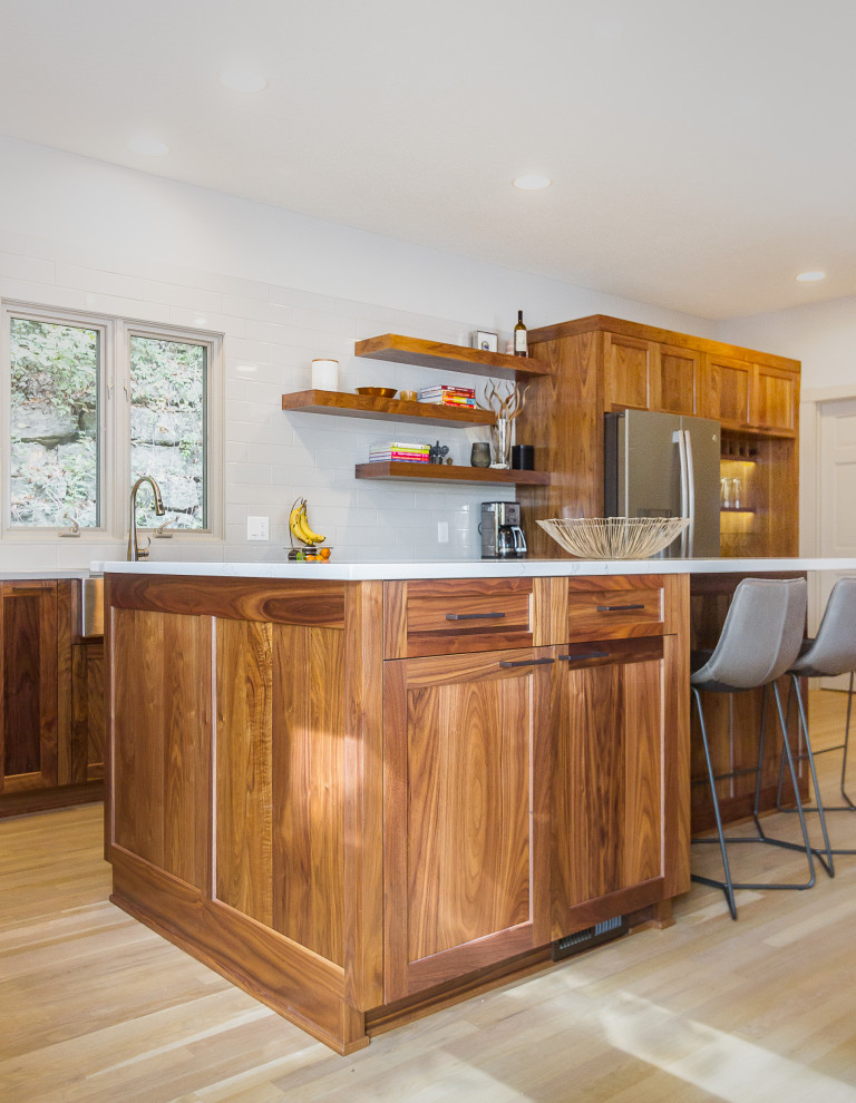 This is an example of a mid-sized contemporary l-shaped eat-in kitchen in Kansas City with a farmhouse sink, recessed-panel cabinets, medium wood cabinets, quartz benchtops, white splashback, ceramic splashback, stainless steel appliances, light hardwood floors, with island and white benchtop.