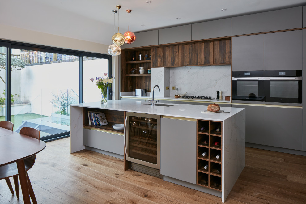 Large contemporary grey and brown l-shaped open plan kitchen in London with a built-in sink, flat-panel cabinets, grey cabinets, quartz worktops, grey splashback, engineered quartz splashback, stainless steel appliances, medium hardwood flooring, an island, grey worktops and feature lighting.