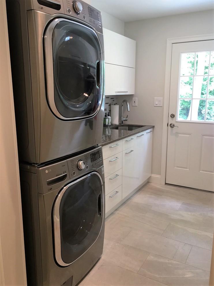This is an example of a small transitional single-wall dedicated laundry room in Philadelphia with an undermount sink, flat-panel cabinets, white cabinets, quartz benchtops, grey walls, light hardwood floors, a stacked washer and dryer, brown floor and grey benchtop.