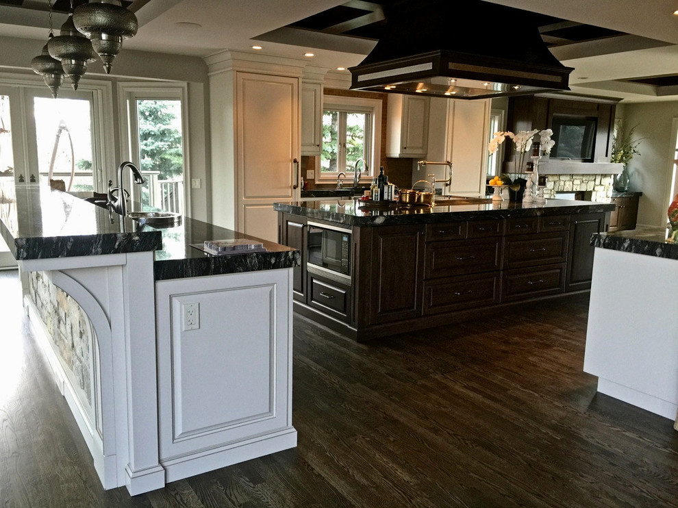 This is an example of a large transitional eat-in kitchen in Calgary with a farmhouse sink, raised-panel cabinets, white cabinets, granite benchtops, beige splashback, stone tile splashback, stainless steel appliances, dark hardwood floors and multiple islands.