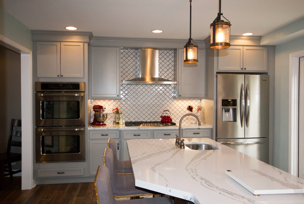 Example of a large transitional l-shaped medium tone wood floor and brown floor enclosed kitchen design in Cincinnati with a farmhouse sink, recessed-panel cabinets, gray cabinets, marble countertops, white backsplash, porcelain backsplash, stainless steel appliances and an island