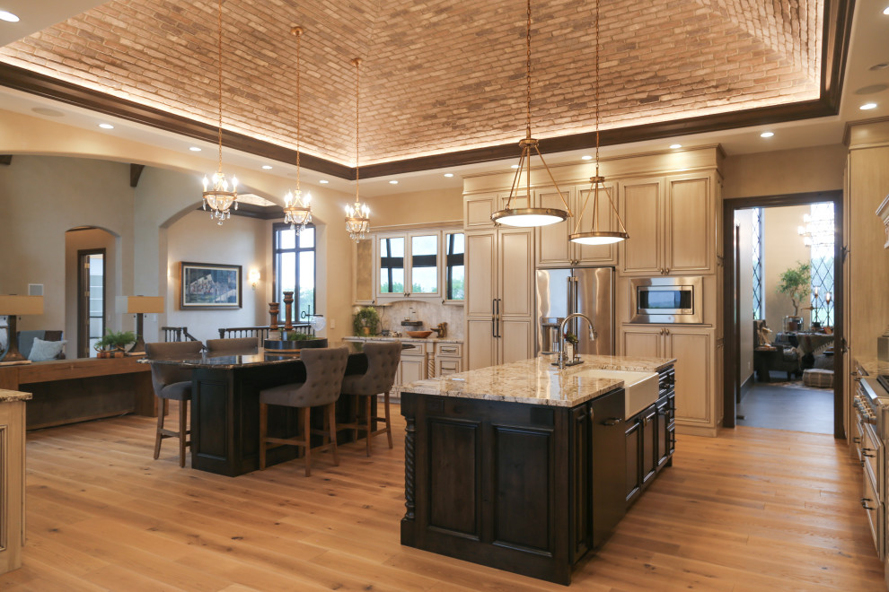 This is an example of a large u-shaped open plan kitchen in Other with a farmhouse sink, raised-panel cabinets, white cabinets, granite benchtops, beige splashback, stone tile splashback, stainless steel appliances, medium hardwood floors, multiple islands, brown floor, multi-coloured benchtop and vaulted.
