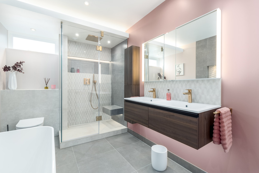На фото: детская ванная комната среднего размера в стиле модернизм с плоскими фасадами, коричневыми фасадами, отдельно стоящей ванной, душевой комнатой, инсталляцией, серой плиткой, керамической плиткой, розовыми стенами, полом из керамогранита, подвесной раковиной, столешницей из искусственного камня, серым полом, душем с распашными дверями, белой столешницей, нишей, тумбой под две раковины, подвесной тумбой и многоуровневым потолком с