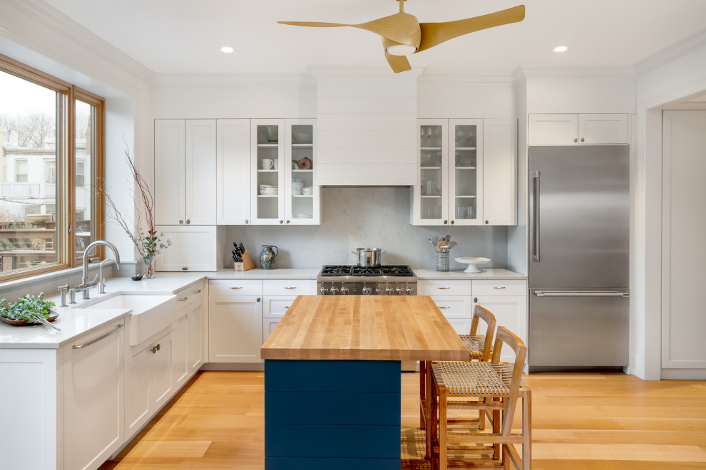 This is an example of a mid-sized transitional l-shaped eat-in kitchen in New York with a farmhouse sink, shaker cabinets, white cabinets, quartz benchtops, grey splashback, stone slab splashback, stainless steel appliances, light hardwood floors, with island and grey benchtop.