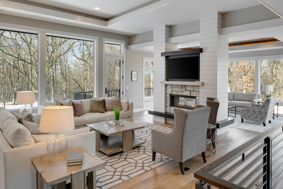 This is an example of a transitional open concept living room in Minneapolis with grey walls, medium hardwood floors, a two-sided fireplace, a stone fireplace surround, a wall-mounted tv, beige floor and recessed.
