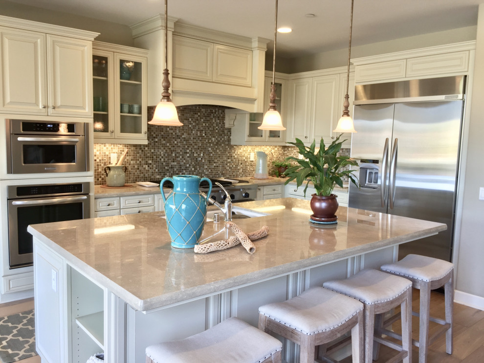 This is an example of a large beach style l-shaped eat-in kitchen in Orange County with a double-bowl sink, recessed-panel cabinets, white cabinets, limestone benchtops, multi-coloured splashback, mosaic tile splashback, stainless steel appliances, light hardwood floors, with island and beige benchtop.