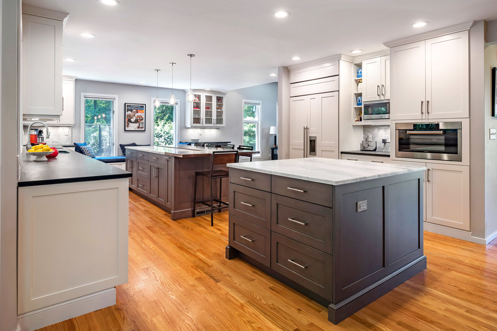 Photo of a large transitional l-shaped eat-in kitchen in Bridgeport with a single-bowl sink, recessed-panel cabinets, white cabinets, marble benchtops, white splashback, marble splashback, stainless steel appliances, medium hardwood floors, multiple islands, brown floor and white benchtop.