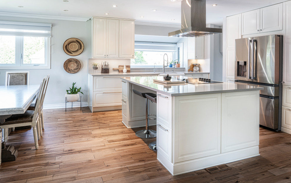 This is an example of a mid-sized contemporary l-shaped eat-in kitchen in Montreal with a farmhouse sink, shaker cabinets, white cabinets, quartz benchtops, grey splashback, engineered quartz splashback, stainless steel appliances, medium hardwood floors, with island, brown floor and grey benchtop.