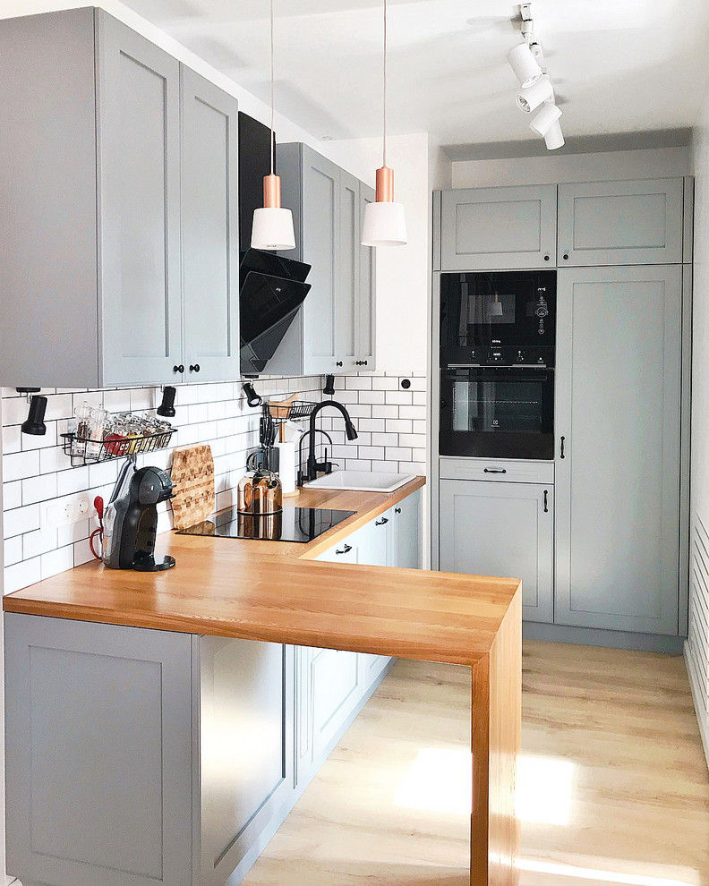 This is an example of a contemporary l-shaped kitchen in Moscow with a drop-in sink, recessed-panel cabinets, grey cabinets, wood benchtops, white splashback, black appliances, no island, brown floor and brown benchtop.