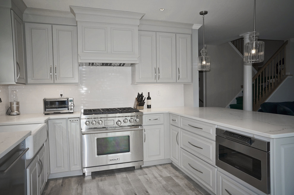 Photo of a mid-sized transitional u-shaped kitchen in New York with a farmhouse sink, raised-panel cabinets, grey cabinets, quartz benchtops, white splashback, subway tile splashback, stainless steel appliances, vinyl floors, a peninsula and grey floor.