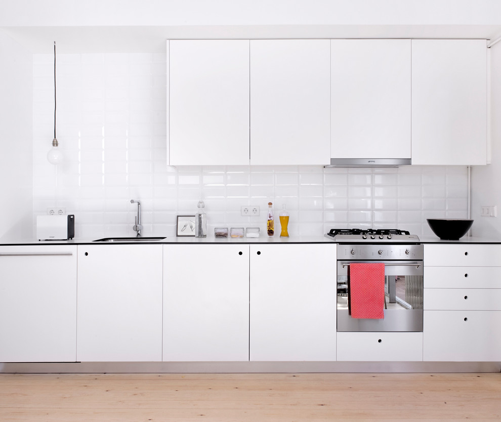 This is an example of a mid-sized industrial single-wall separate kitchen in Barcelona with an undermount sink, flat-panel cabinets, white cabinets, white splashback, subway tile splashback, stainless steel appliances, light hardwood floors and no island.