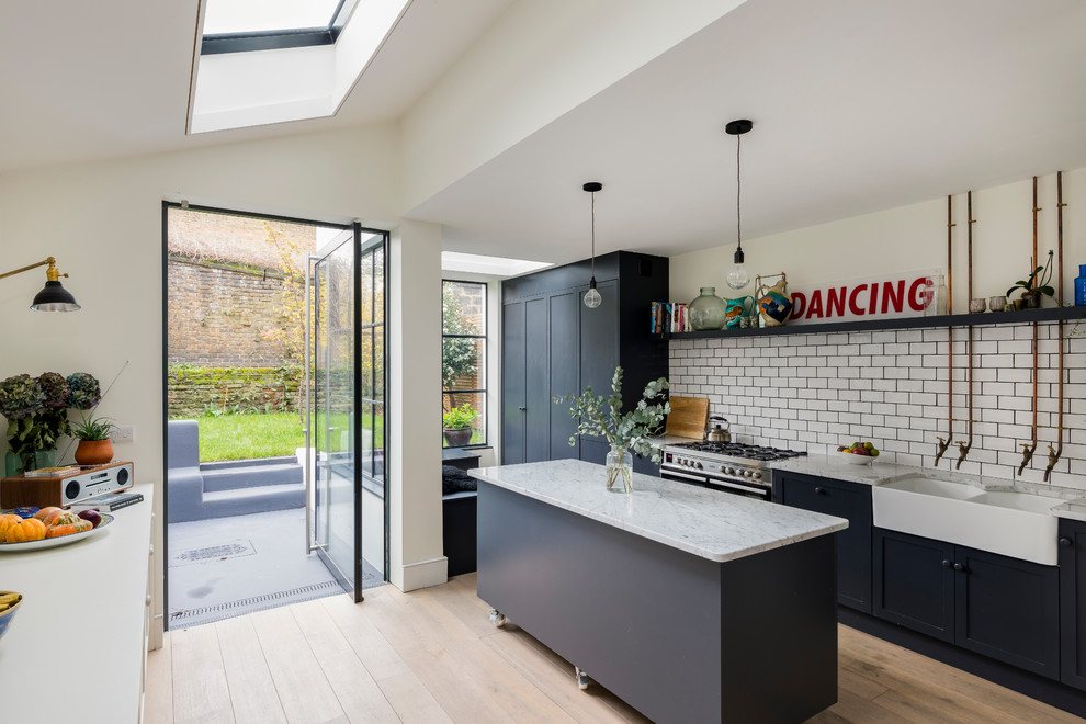 Inspiration for a contemporary kitchen in London with a farmhouse sink, shaker cabinets, black cabinets, white splashback, subway tile splashback, stainless steel appliances, light hardwood floors, with island and beige floor.
