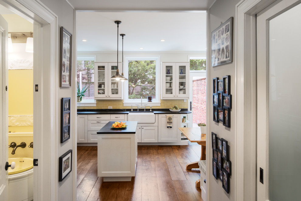 This is an example of a traditional single-wall eat-in kitchen in New York with a farmhouse sink, shaker cabinets, white cabinets, yellow splashback, subway tile splashback, medium hardwood floors, with island, brown floor and black benchtop.