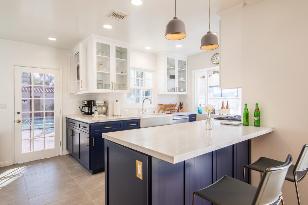 Mid-sized transitional u-shaped open plan kitchen in Los Angeles with a farmhouse sink, shaker cabinets, blue cabinets, stainless steel appliances, a peninsula, beige floor, beige benchtop, marble benchtops and porcelain floors.