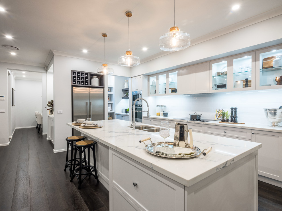 This is an example of a contemporary galley open plan kitchen in Hobart with a double-bowl sink, shaker cabinets, white cabinets, marble benchtops, white splashback, ceramic splashback, stainless steel appliances, medium hardwood floors, with island, brown floor and white benchtop.