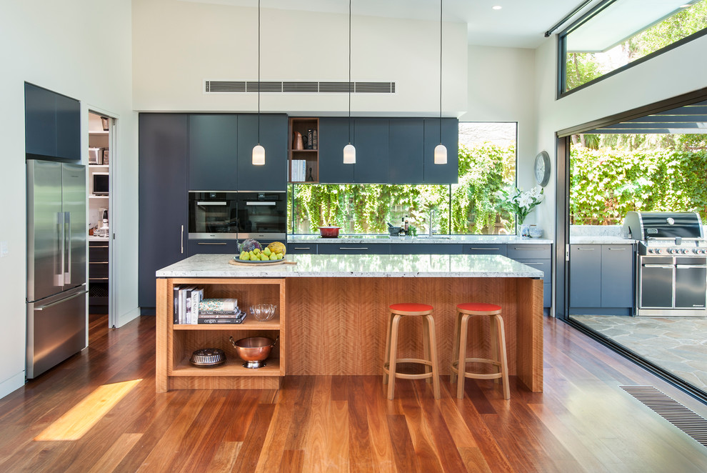 This is an example of a large transitional single-wall open plan kitchen in Melbourne with a double-bowl sink, blue cabinets, granite benchtops, black appliances, medium hardwood floors, with island, white benchtop and window splashback.