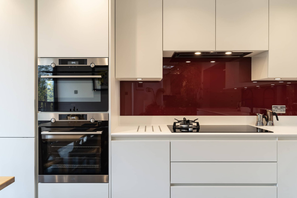 Mid-sized traditional l-shaped eat-in kitchen in Oxfordshire with an integrated sink, white cabinets, red splashback, glass sheet splashback, stainless steel appliances, ceramic floors, no island, black floor and white benchtop.