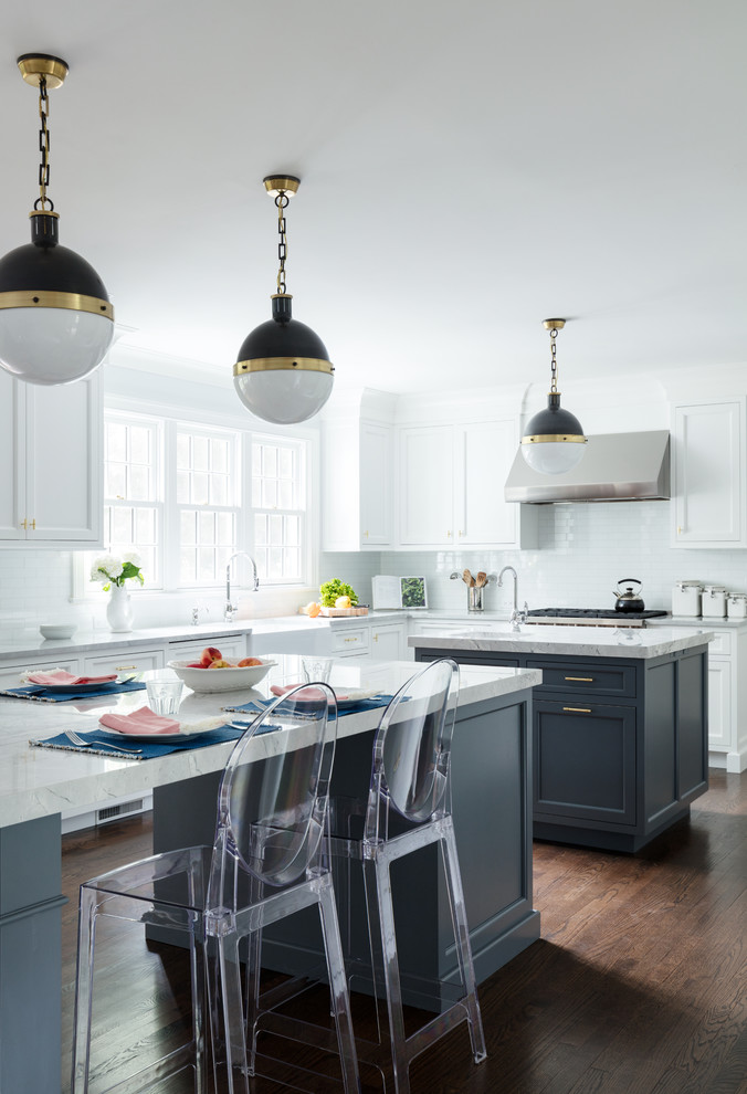 Photo of a transitional l-shaped kitchen in New York with a farmhouse sink, marble benchtops, white splashback, multiple islands, brown floor, dark hardwood floors, shaker cabinets, white cabinets, subway tile splashback and grey benchtop.