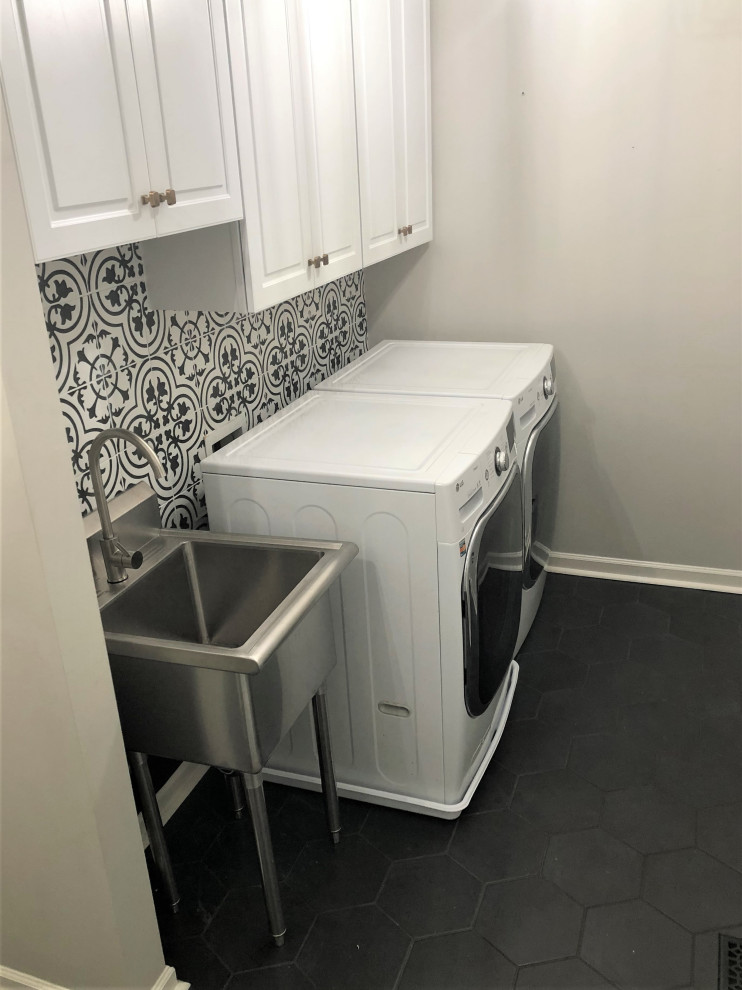 Photo of a small galley dedicated laundry room in DC Metro with an utility sink, recessed-panel cabinets, white cabinets, porcelain splashback, grey walls, porcelain floors, a side-by-side washer and dryer and black floor.