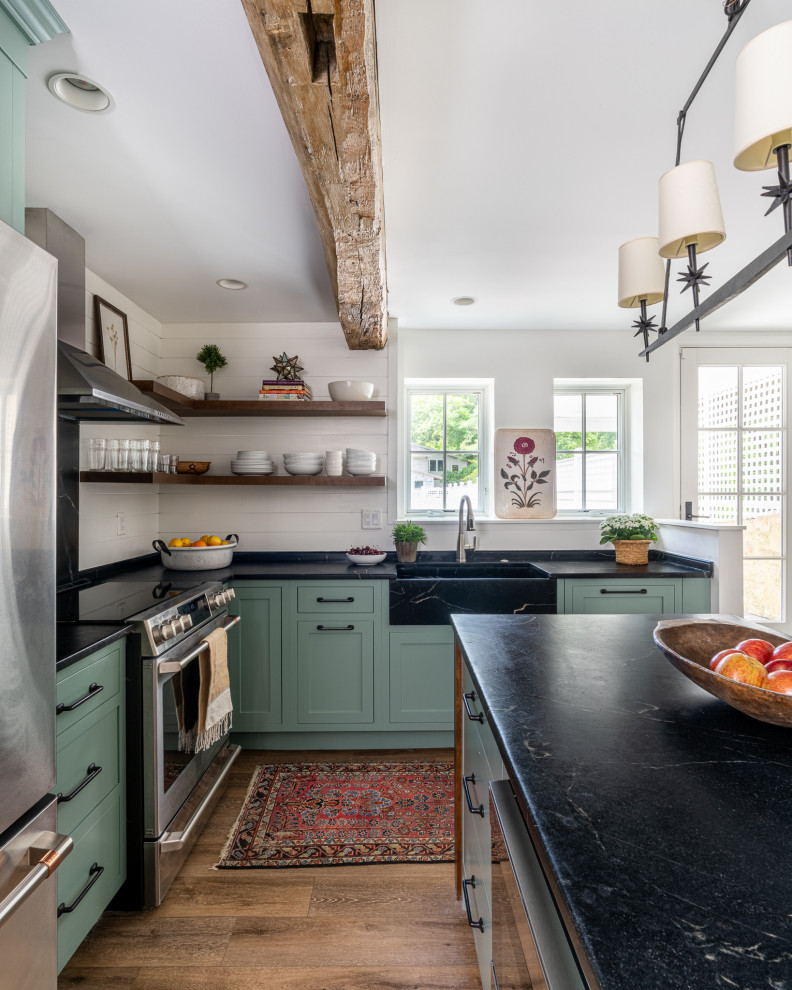 This is an example of a small rural l-shaped enclosed kitchen in Philadelphia with a belfast sink, shaker cabinets, green cabinets, soapstone worktops, black splashback, stone slab splashback, stainless steel appliances, laminate floors, an island, brown floors and black worktops.