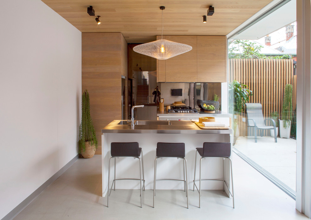 This is an example of a small contemporary galley kitchen in Melbourne with an integrated sink, flat-panel cabinets, light wood cabinets, stainless steel benchtops, stainless steel appliances, concrete floors, with island, grey floor and metallic splashback.