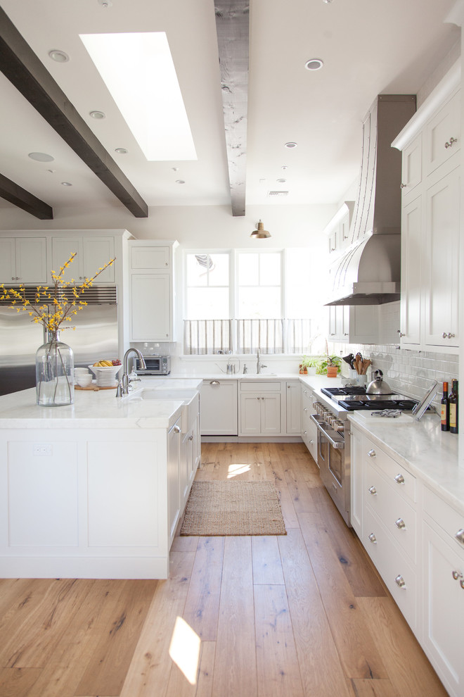 Large transitional l-shaped light wood floor eat-in kitchen photo in Phoenix with a farmhouse sink, white cabinets, white backsplash, stainless steel appliances, an island, shaker cabinets, stainless steel countertops and porcelain backsplash