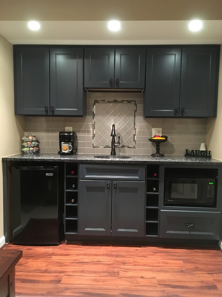 This is an example of a mid-sized transitional galley wet bar in DC Metro with an undermount sink, recessed-panel cabinets, blue cabinets, granite benchtops, grey splashback, porcelain splashback, vinyl floors and brown floor.
