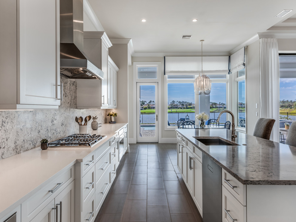 Inspiration for a mid-sized beach style l-shaped eat-in kitchen in Miami with a drop-in sink, raised-panel cabinets, white cabinets, granite benchtops, grey splashback, stone tile splashback, stainless steel appliances, light hardwood floors, with island, grey floor and grey benchtop.