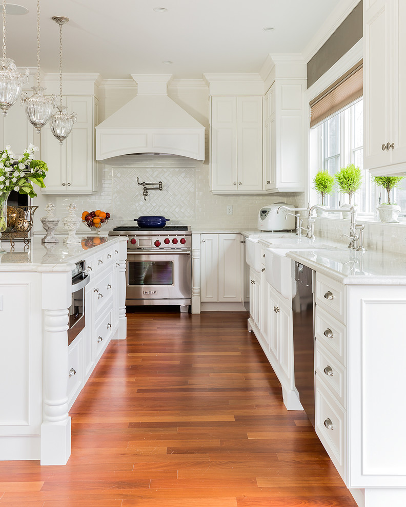 Large traditional kitchen in Boston with a farmhouse sink, white cabinets, quartzite benchtops, white splashback, stainless steel appliances, medium hardwood floors, with island and recessed-panel cabinets.