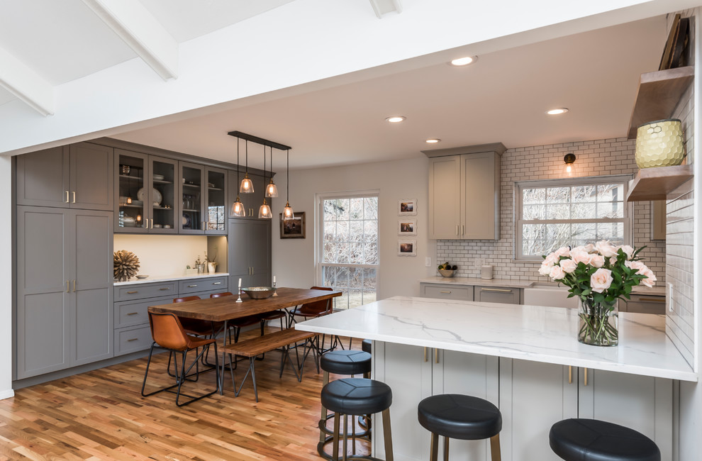 Example of a mid-sized transitional galley medium tone wood floor eat-in kitchen design in Denver with a farmhouse sink, shaker cabinets, gray cabinets, quartz countertops, white backsplash, porcelain backsplash, stainless steel appliances, a peninsula and white countertops