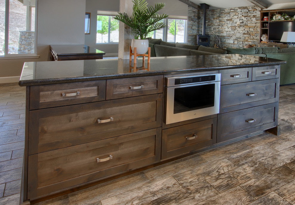 Large mountain style l-shaped porcelain tile, brown floor and vaulted ceiling open concept kitchen photo with an undermount sink, shaker cabinets, brown cabinets, granite countertops, brown backsplash, marble backsplash, stainless steel appliances, an island and black countertops