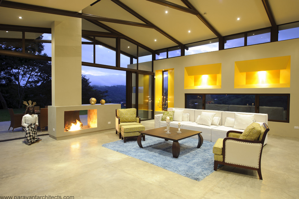 Cette image montre un grand salon design ouvert avec un mur jaune, sol en béton ciré, une cheminée double-face, un manteau de cheminée en béton, un sol gris et poutres apparentes.