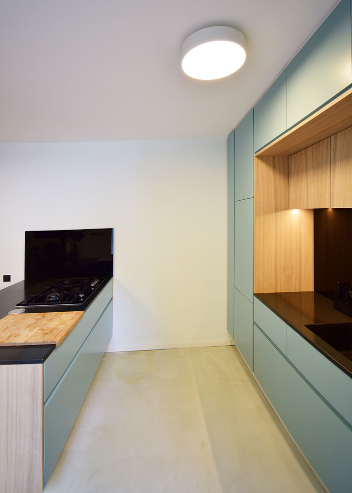 Design ideas for a mid-sized modern galley open plan kitchen in Cologne with an undermount sink, flat-panel cabinets, turquoise cabinets, black splashback, glass sheet splashback, black appliances, concrete floors, with island, grey floor and black benchtop.