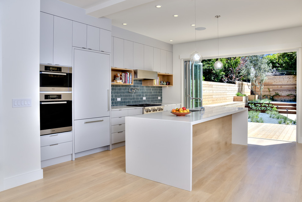This is an example of a mid-sized modern single-wall open plan kitchen in San Francisco with an undermount sink, flat-panel cabinets, grey cabinets, solid surface benchtops, blue splashback, ceramic splashback, panelled appliances, light hardwood floors and with island.