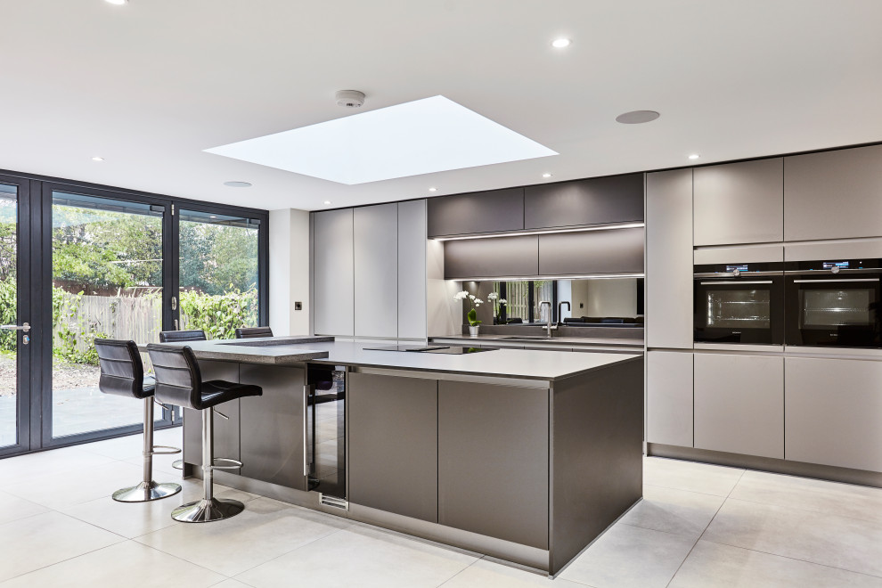 Design ideas for a mid-sized contemporary single-wall open plan kitchen in Other with an integrated sink, flat-panel cabinets, grey cabinets, quartzite benchtops, metallic splashback, mirror splashback, black appliances, porcelain floors, with island, grey floor, grey benchtop and vaulted.