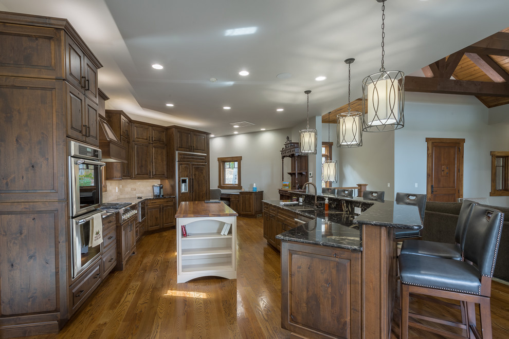 Photo of a large country l-shaped open plan kitchen in Charlotte with a double-bowl sink, raised-panel cabinets, dark wood cabinets, beige splashback, stainless steel appliances, medium hardwood floors, multiple islands and brown floor.