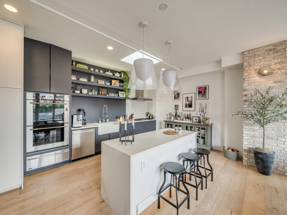 Photo of an industrial kitchen in New York with a farmhouse sink, flat-panel cabinets, grey cabinets, grey splashback, stainless steel appliances, light hardwood floors, with island, beige floor and white benchtop.