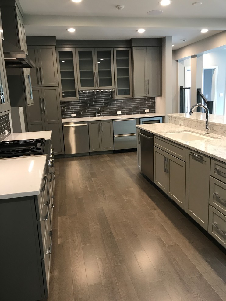 Mid-sized transitional l-shaped open plan kitchen in St Louis with an undermount sink, recessed-panel cabinets, grey cabinets, quartz benchtops, black splashback, ceramic splashback, stainless steel appliances, medium hardwood floors, with island and brown floor.
